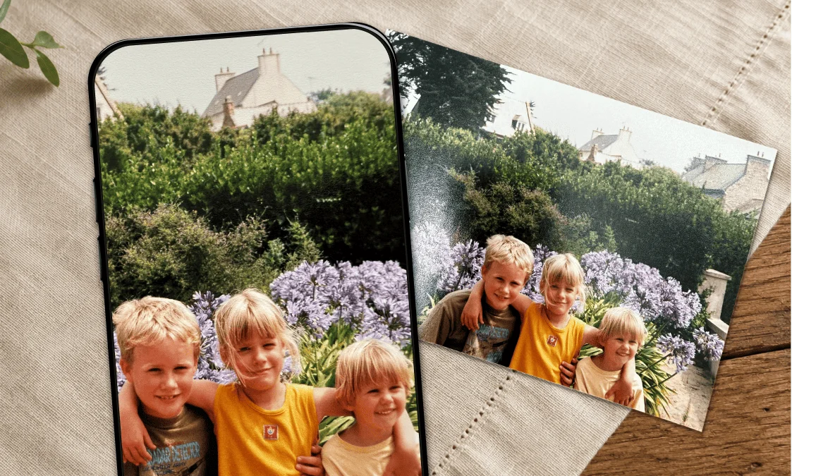 A faded printed photo of three children in a garden lying on a linen cloth, next to a phone showing the same photo restored with brighter colour and sharper detail